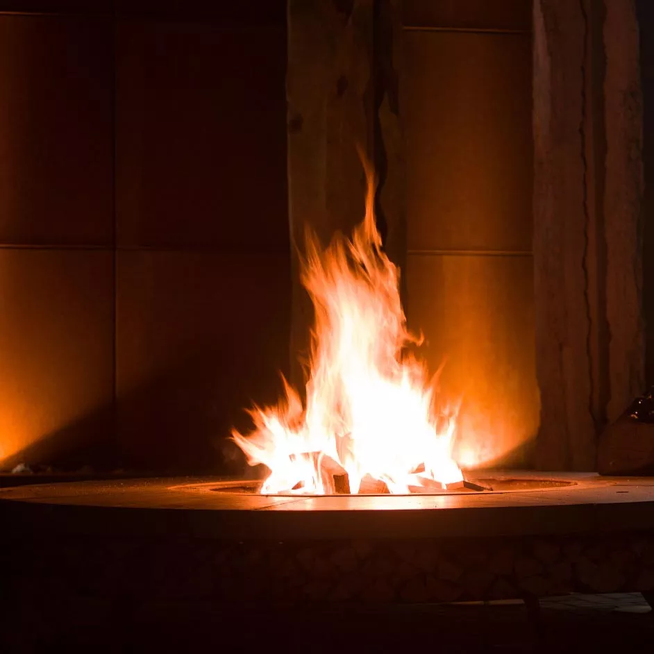 AK47 Ercole Beton Stahl Feuerstelle in Betrieb mit lodernden Flammen im Hintergrund stimmungsvolle Beleuchtung und gemütliche Atmosphäre