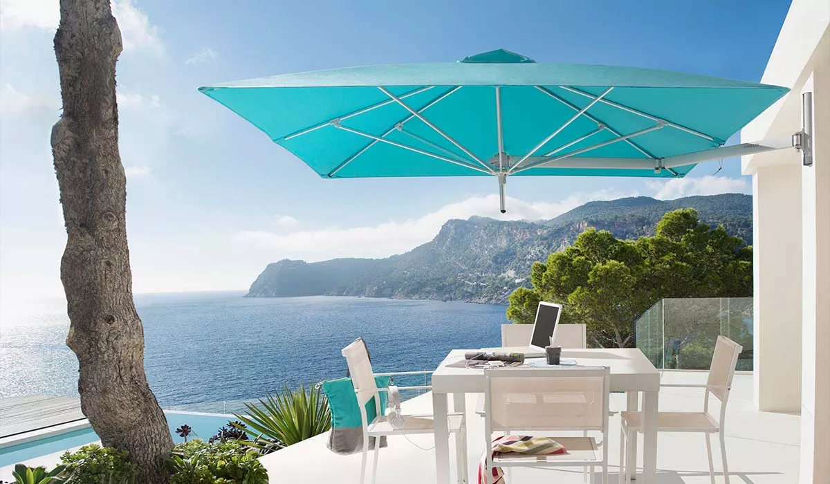 Paraflex Wandsonnenschirm in Türkis über einem weißen Terrassentisch mit Meerblick im Hintergrund.