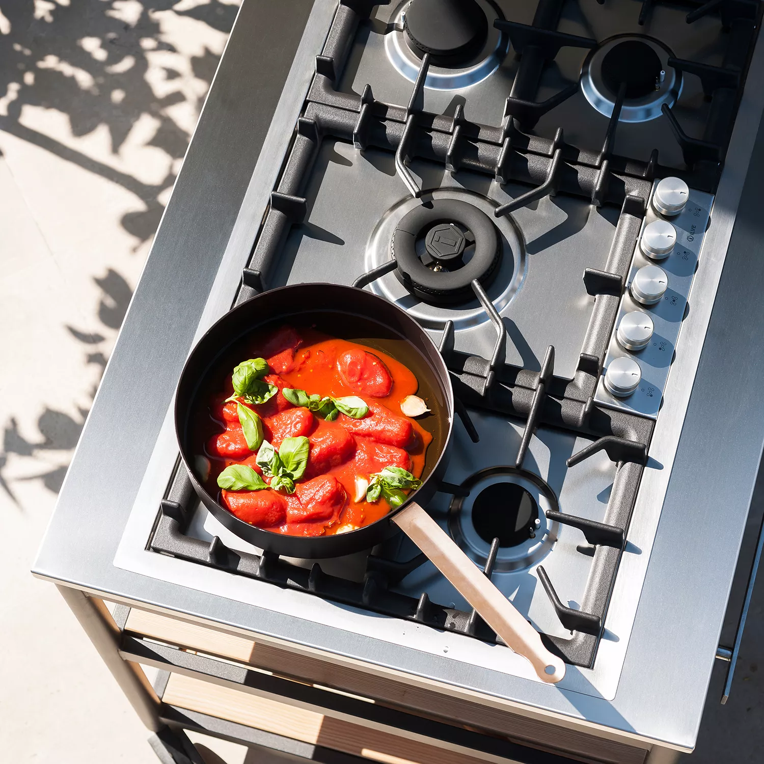 Roda Norma Küchenmodul in Edelstahl mit Gaskochfeld, darauf eine Pfanne mit Tomaten und Basilikum, von oben fotografiert.