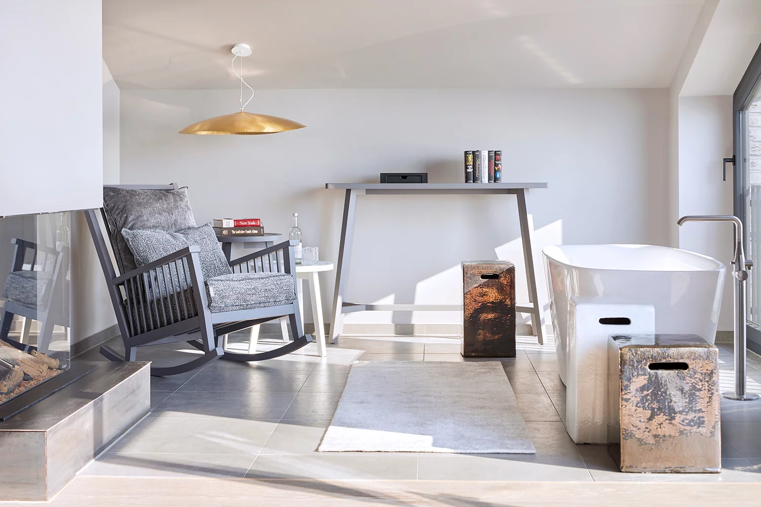 Ein modernes Badezimmer mit Gervasoni Suite Möbeln darunter ein Schaukelstuhl ein Tisch und eine Badewanne in einem hellen Raum mit großen Fenstern.