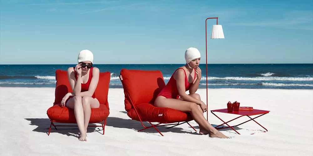 Zwei Frauen in roten Diabla Valentina Sesseln am Strand, mit rotem Beistelltisch und Lampe, vor blauem Himmel und Meer.