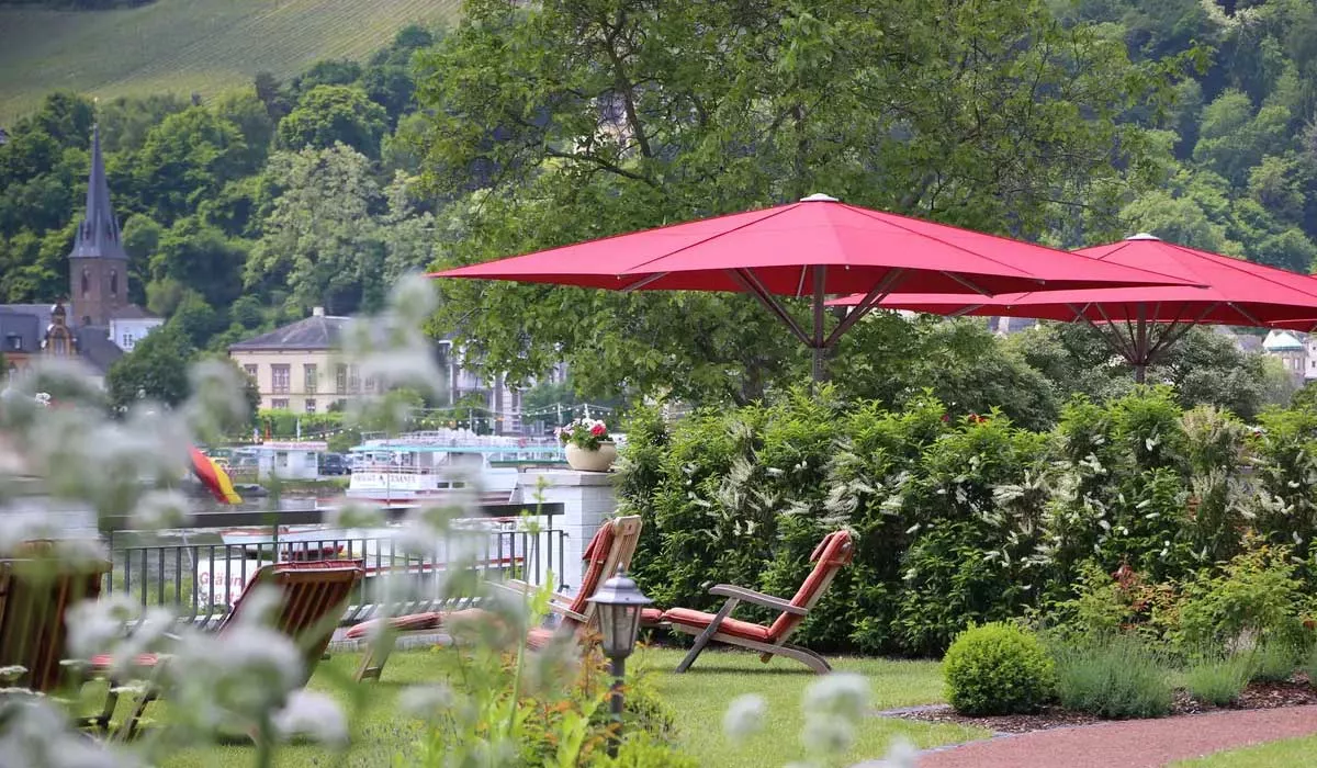 Rote May Sonnenschirme Filius spenden Schatten in einem idyllischen Garten mit Blick auf einen Fluss und eine Kirche im Hintergrund.