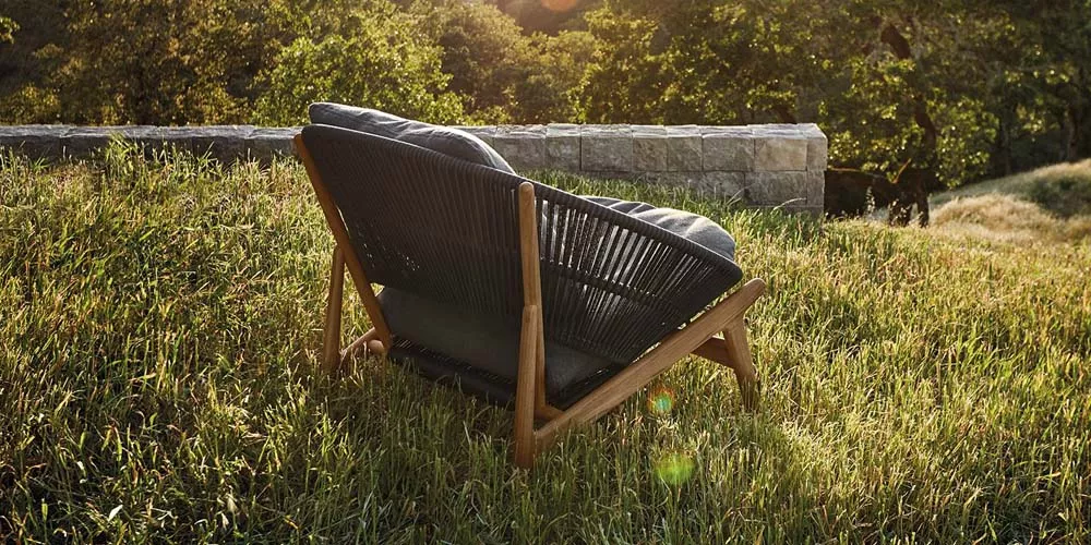 Gartenstuhl von Gloster, Modell Bora, in Schwarz mit Holzrahmen, auf einer Wiese vor einer Steinmauer bei Sonnenuntergang.