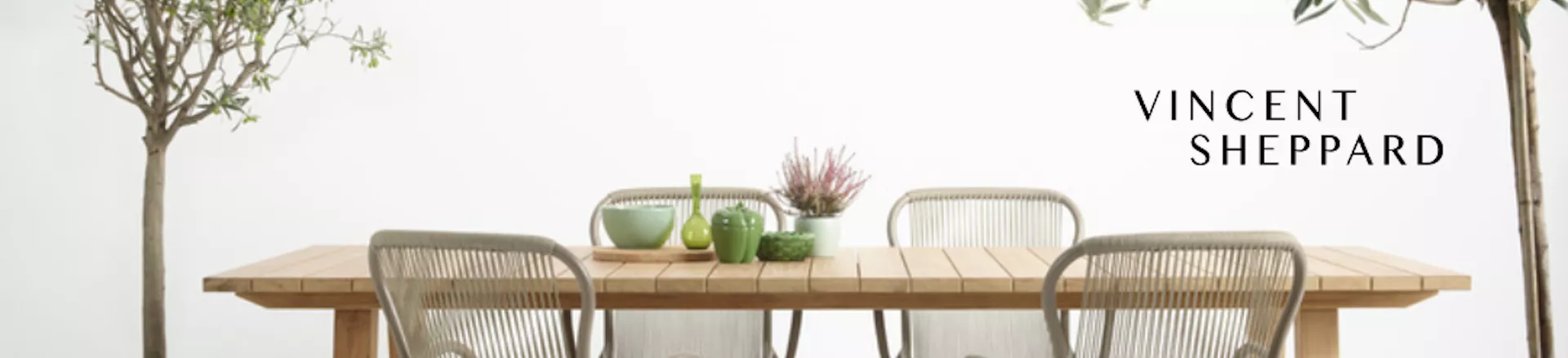 Vincent Sheppard Banner mit Holztisch und grauen Stühlen vor weißem Hintergrund, dekoriert mit grünen und violetten Accessoires.