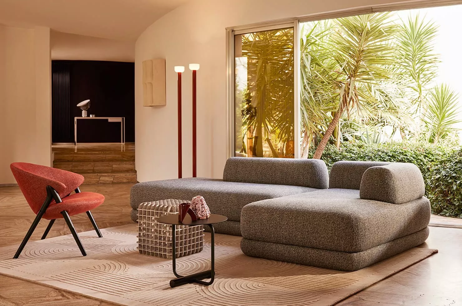 Ein Wohnzimmer mit einem Zanotta Sofa und einem roten Sessel in einer modernen Einrichtung mit großen Fenstern und Pflanzen im Hintergrund.