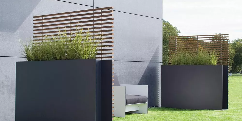 Conmoto Sotomon Pflanzkübel in Anthrazit mit Holzlamellen, auf einer Terrasse vor einer grauen Wand, im Hintergrund grüner Rasen.