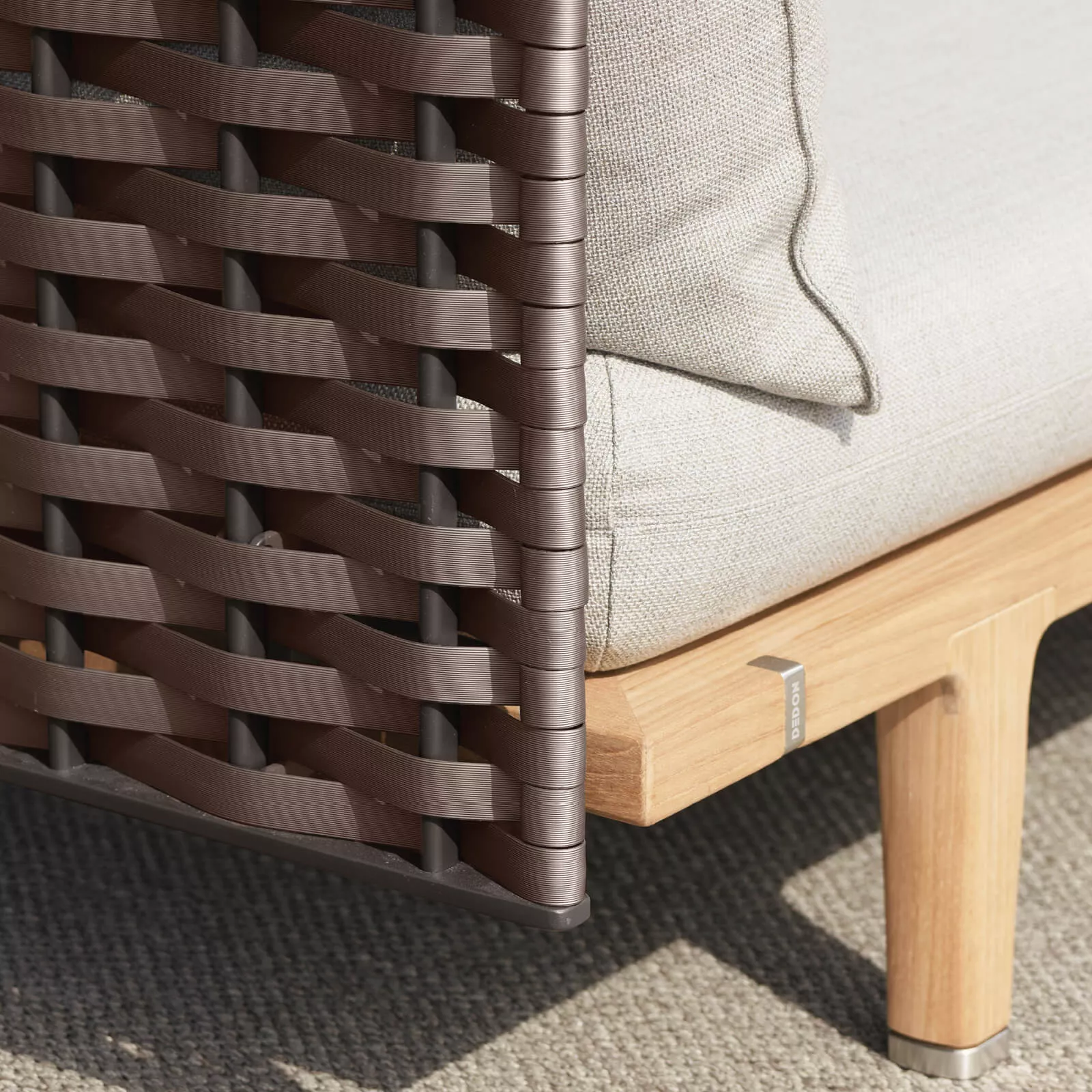 Dedon Sealine Lounge zeigt eine Nahaufnahme eines geflochtenen Seitenteils und eines Teakholzrahmens mit einem beigen Kissen auf einem Teppich.