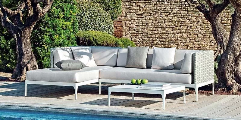 Ethimo Infinity Outdoor Loungegruppe in hellem Grau auf Holzterrasse vor Steinmauer und Bäumen, mit Kissen und Couchtisch, Pool im Vordergrund.