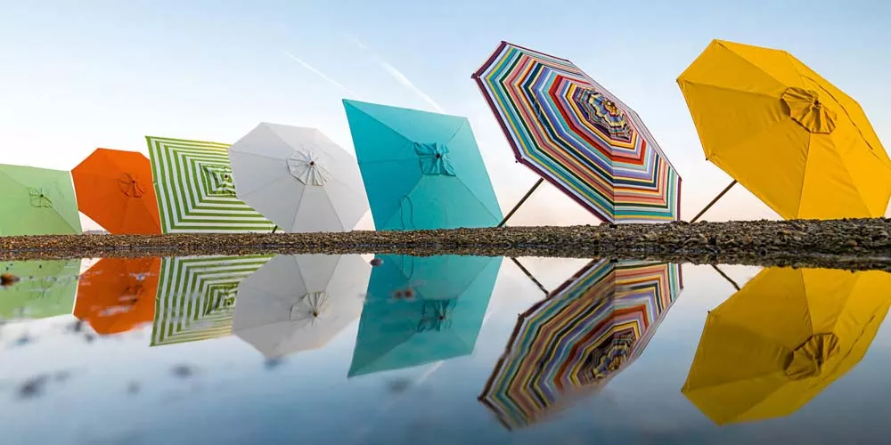 Bunte Weishäupl Klassiker Sonnenschirme in verschiedenen Farben spiegeln sich in einer Wasserfläche vor klarem Himmel.