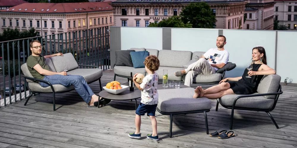 Vier Personen entspannen auf grauen Todus Starling Sofas auf einer Dachterrasse mit Holzboden und Stadtblick bei Abenddämmerung.