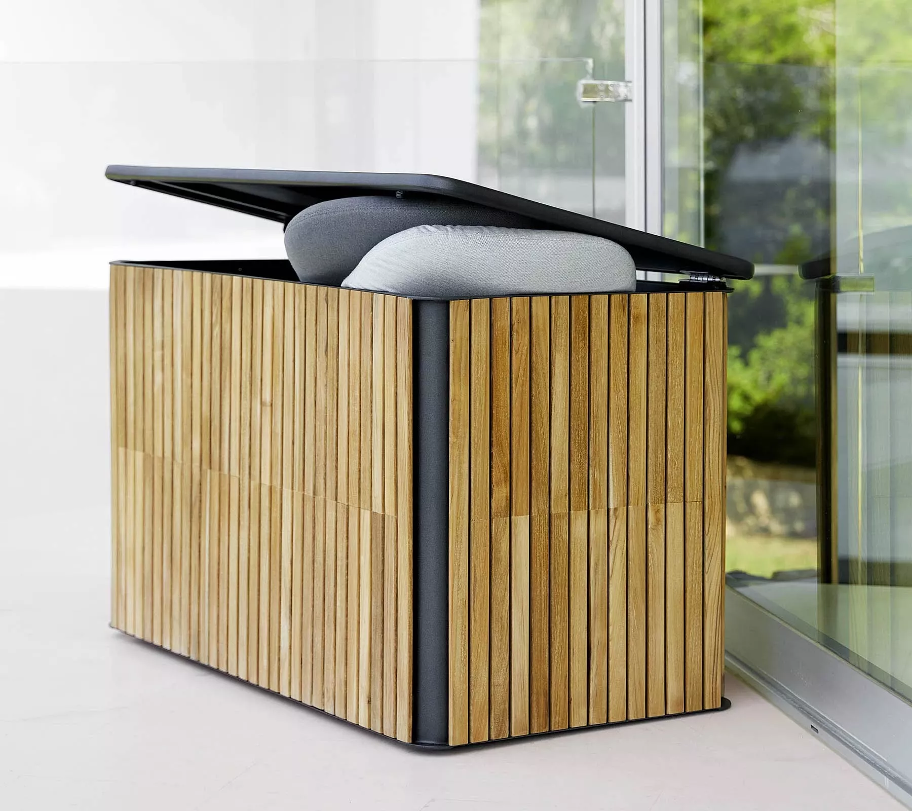 Cane-line Combine Kissentruhe in Teak, geöffnet, zeigt graue Kissen, auf einer Terrasse mit Glaswand im Hintergrund.
