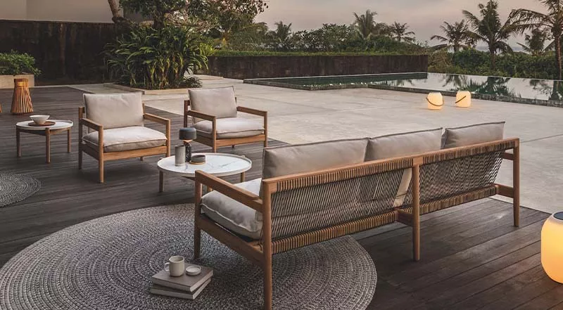 Gloster Sanac Lounge-Set aus Holz und Stoff in Naturtönen auf einer Terrasse mit Pool und Palmen im Hintergrund bei Sonnenuntergang.