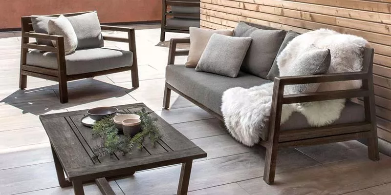 Ethimo Costes Outdoor Loungegruppe in dunklem Holz mit grauen Kissen auf einer Terrasse mit Holzwand im Hintergrund.