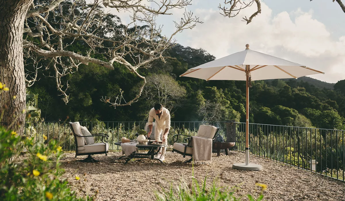 Ein weißer Glatz Sonnenschirm steht auf einer Terrasse mit Gartenmöbeln, umgeben von grüner Natur und einem bewölkten Himmel.
