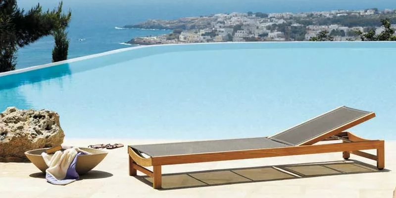 Ethimo Sand Outdoor-Sonnenliege in Beige auf einer Terrasse mit Meerblick und Infinity-Pool im Hintergrund.