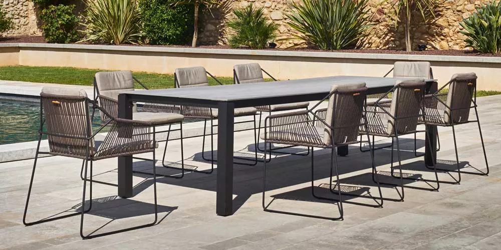 Oasiq Machar Essgruppe in Grau auf einer Terrasse, bestehend aus einem großen Tisch und acht Stühlen, vor einer Steinmauer im Garten.