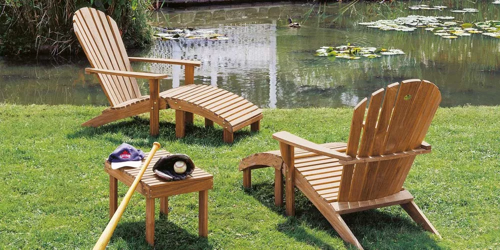 Barlow Tyrie Adirondack Loungegruppe aus Holz auf einer grünen Wiese vor einem Teich, bestehend aus zwei Stühlen, einem Tisch und einer Fußbank.