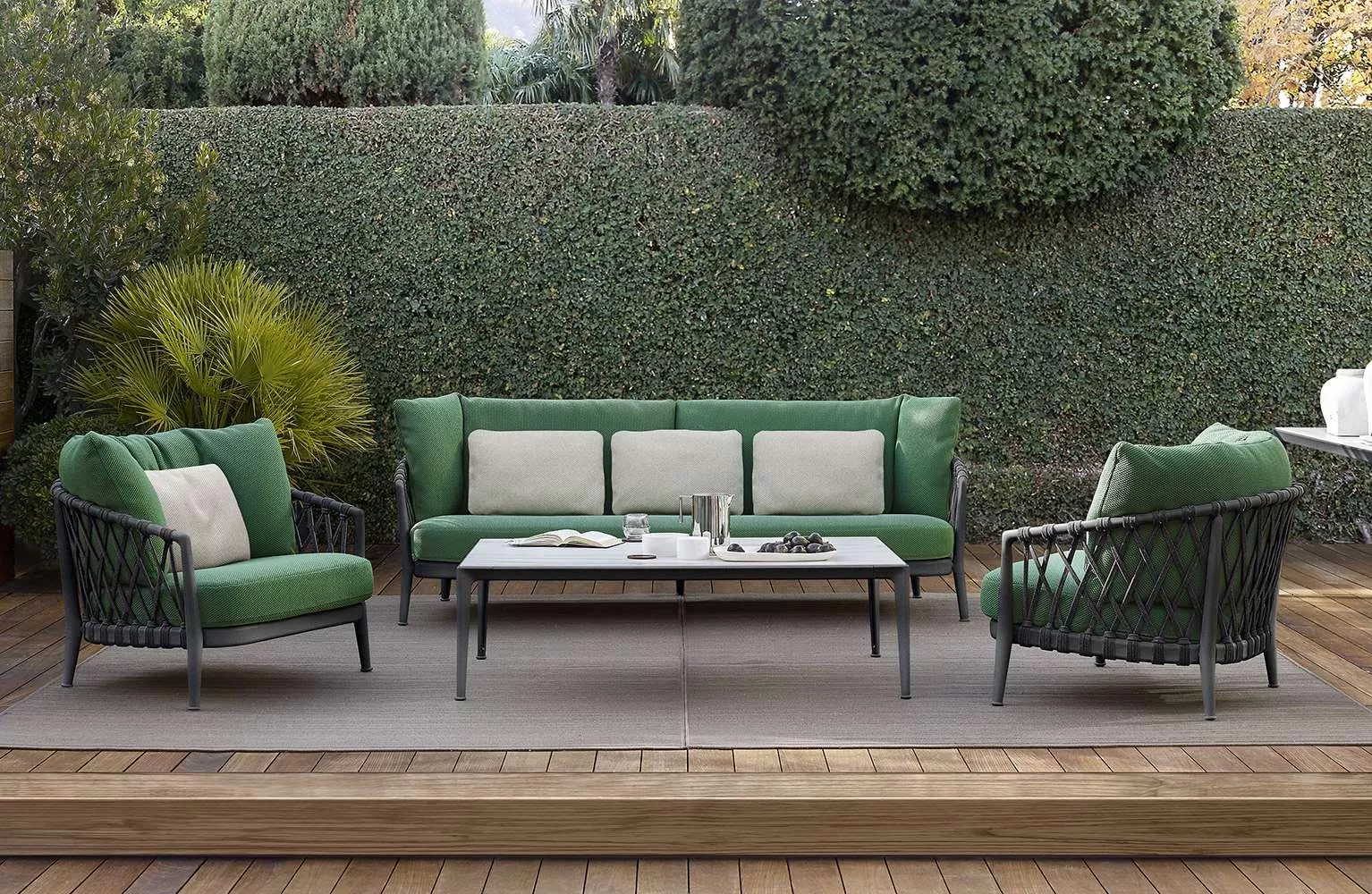 B&B Italia Erica Loungesofa in Grün auf einer Holzterrasse vor einer Hecke, umgeben von passenden Sesseln und einem Tisch.