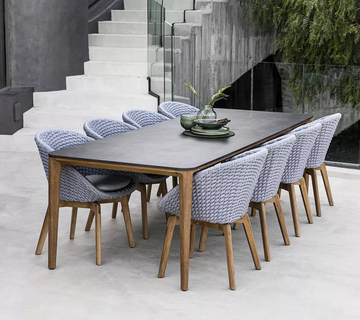 2er-Set Cane-line Peacock Soft Rope Gartenstuhl in Grau mit Teakholz-Beinen, auf modernem Betonboden vor einer Treppe arrangiert.