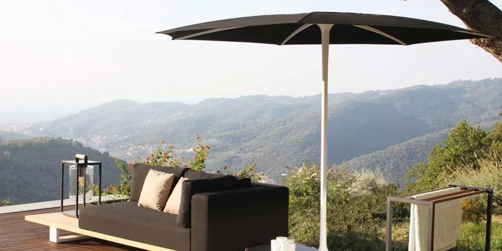 Schwarzer Royal Botania Palma Sonnenschirm auf Holzterrasse mit Bergblick, neben schwarzem Sofa und Beistelltisch.