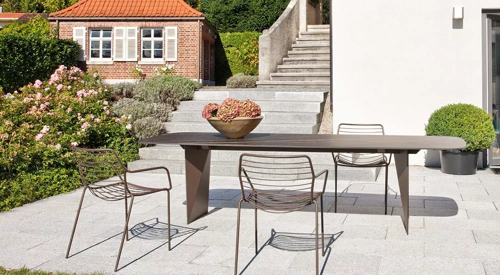 Fischer Möbel Claris Essgruppe in Braun auf einer Terrasse mit Blumen und Treppe im Hintergrund, bestehend aus Tisch und drei Stühlen.