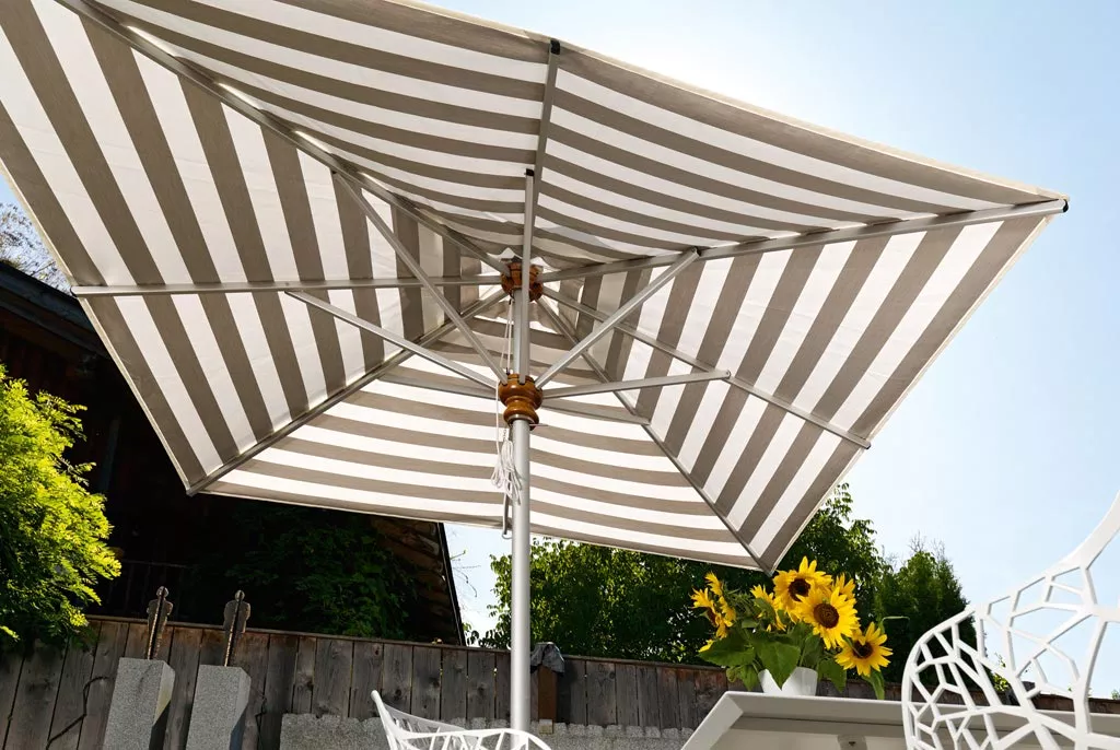 Aluschirm von Weishäupl in Weiß und Grau gestreift, quadratisch, auf einer Terrasse mit Sonnenblumen im Vordergrund.