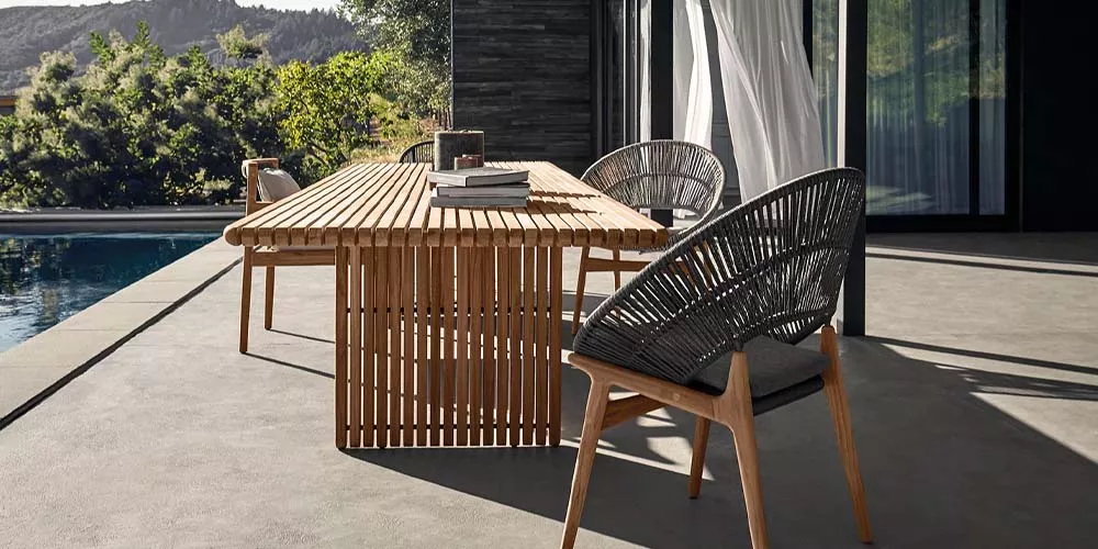 Gloster Deck Kollektion, Holztisch mit grauen Stühlen auf einer Terrasse, im Hintergrund Natur und modernes Gebäude.