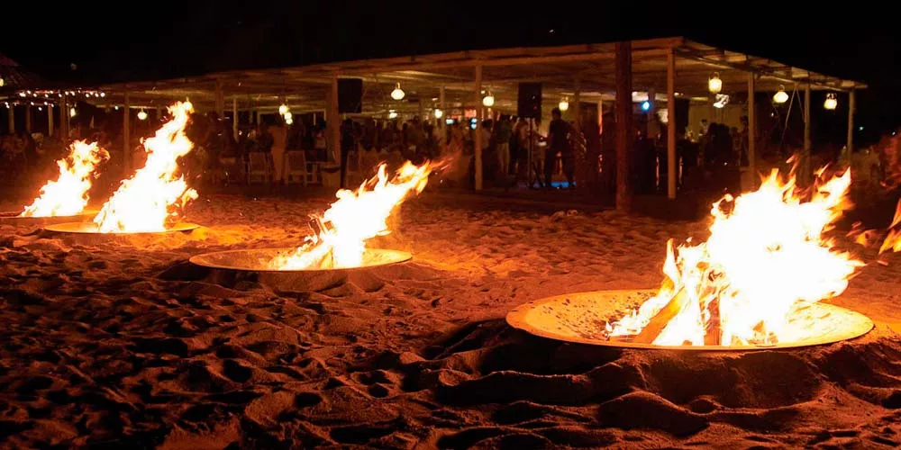 Drei Ak-47 Feuerstellen brennen auf einem sandigen Strand bei Nacht im Hintergrund eine beleuchtete Veranstaltung mit Menschen