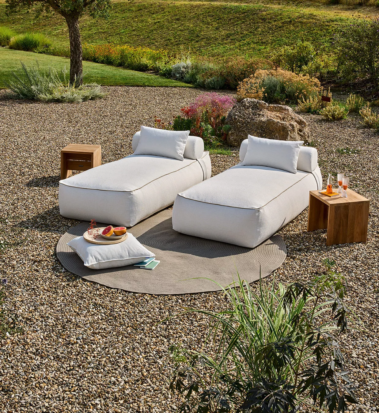 Zwei Maliha Lounge-Betten in Creme mit Kissen, Cabin Beistelltisch 40x40 und Teppich 200 in Taupe auf Kies im Garten.