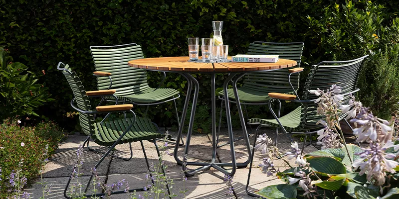 Grüne Houe Click Stühle und runder Holztisch auf einer Terrasse im Garten, umgeben von Pflanzen, bei sonnigem Wetter.