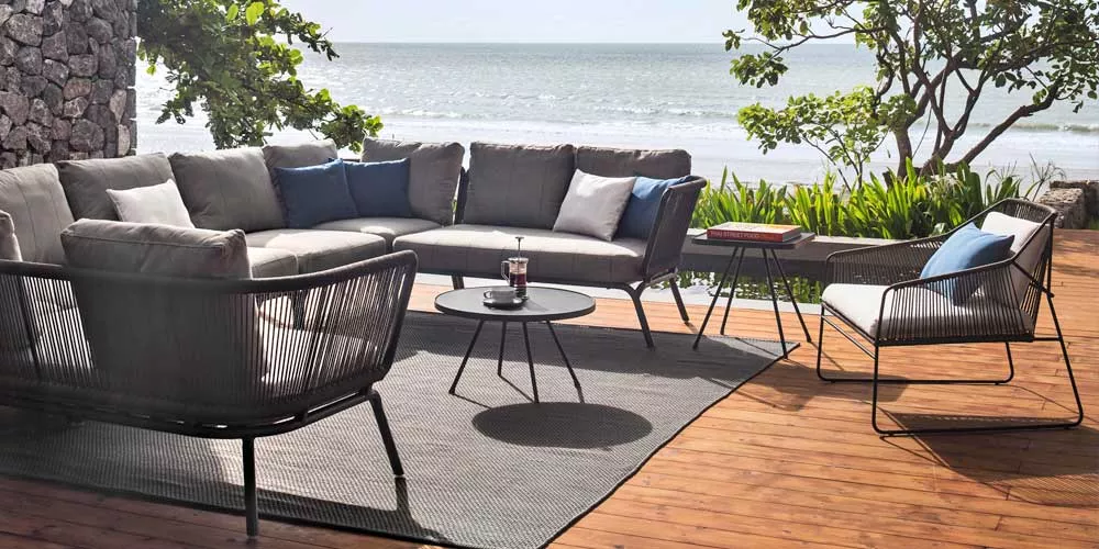 Oasiq Yland Loungegruppe in Grau auf einer Holzterrasse mit Meerblick, bestehend aus einem Ecksofa, Sessel und zwei runden Tischen.