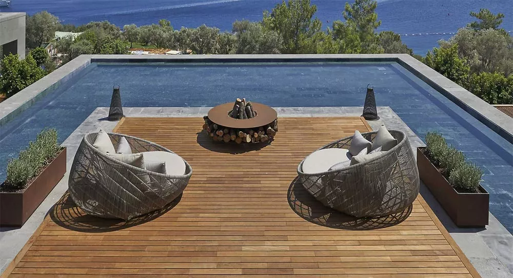 Zwei moderne Sessel stehen auf einer Holzterrasse vor einem Pool mit Blick auf das Meer in der Mitte steht die AK47 Zero Feuerstelle 100 cm Gartenkamin.