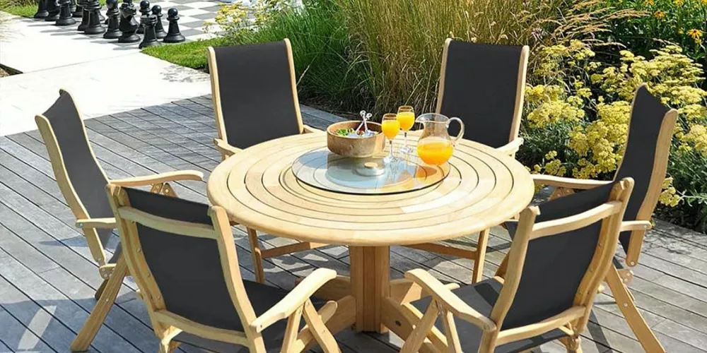 Runder Alexander Rose Roble Gartentisch mit sechs schwarzen Stühlen auf Holzterrasse, umgeben von blühenden Pflanzen, im Freien.