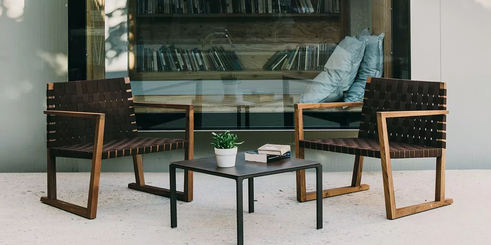 Andreu World Serena Stühle aus Teakholz in einem modernen Außenbereich vor einer Glaswand mit Kissen und einem kleinen Tisch.