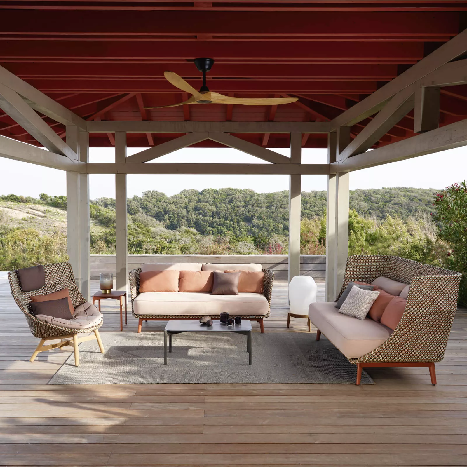 Dedon Mbarq 3er Sofa in einer gemütlichen Outdoor-Szene auf einer Holzterrasse mit Blick auf eine grüne Landschaft und einem Pavillon im Hintergrund