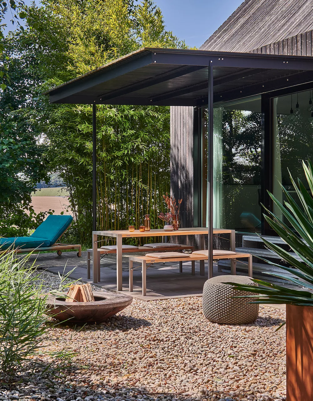 Gartenmöbel-Set Flip in Taupe mit Bank 160x43, Tisch 180x90 und Pouf, auf einer Terrasse mit Kiesboden und grüner Pflanzenkulisse.