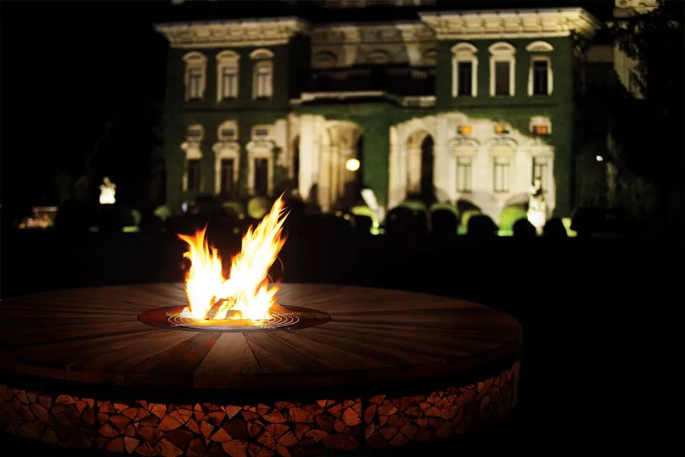 AK47 Art Feuerstelle aus Kastanienholz und Stahl im Garten eines beleuchteten Herrenhauses bei Nacht