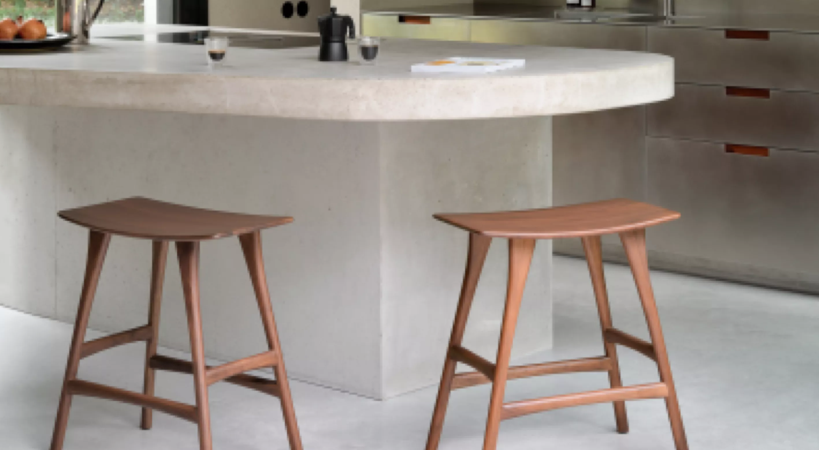 Zwei hölzerne Hocker von Villa Schmidt in einem modernen Küchenambiente mit Betoninsel und Edelstahlgeräten im Hintergrund.