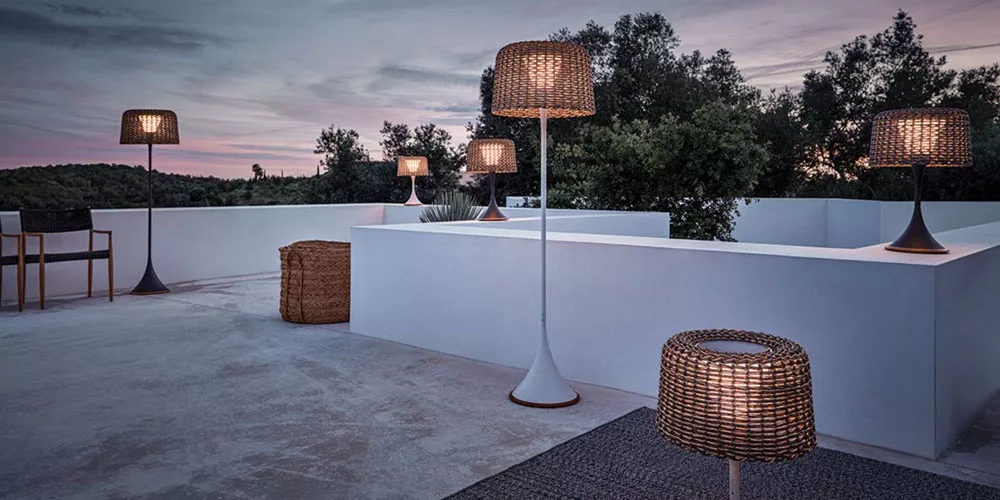 Mehrere braune Kettal Boma Stehleuchten auf einer modernen Terrasse bei Sonnenuntergang, umgeben von Bäumen und weißen Wänden.