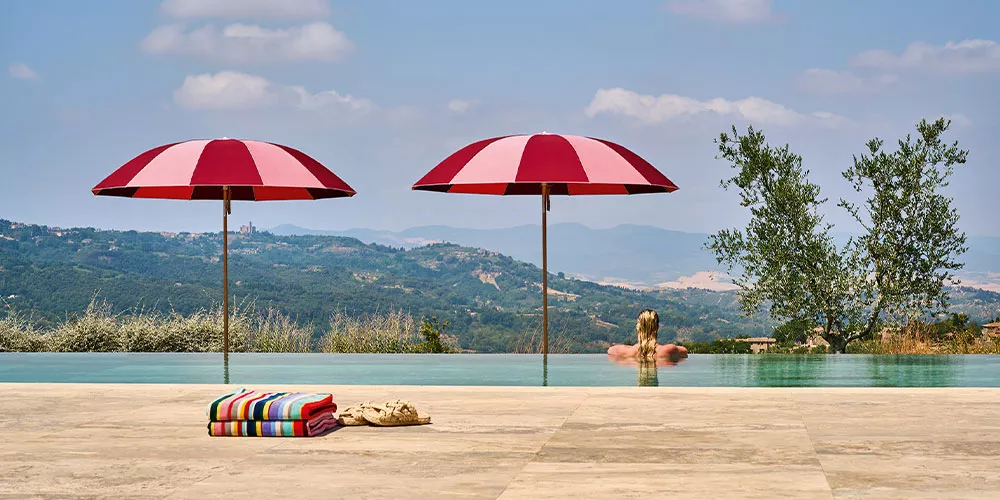Zwei rot-weiße Weishäupl Pagodenschirme am Pool mit Blick auf die Hügel, im Vordergrund bunte Handtücher und ein Strohhut.