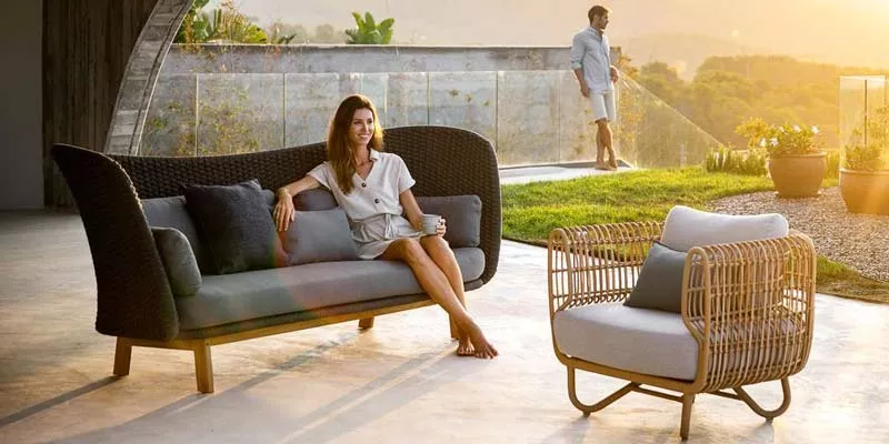 Cane-line Peacock Loungegruppe in Schwarz und Natur auf einer Terrasse bei Sonnenuntergang, mit entspannter Frau auf dem Sofa.
