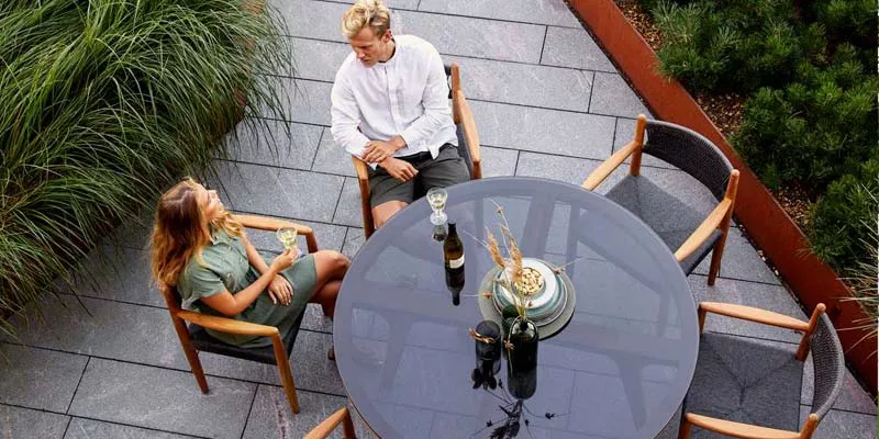 Zwei Personen sitzen an einem runden, schwarzen Cane-line Royal Esstisch im Freien, umgeben von grünen Pflanzen.