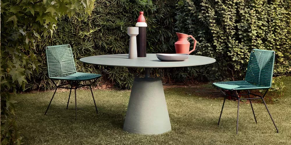 Grüner MDF Italia Neil Twist Tisch mit zwei Stühlen im Garten, umgeben von Pflanzen, als stimmungsvolles Outdoor-Setting.