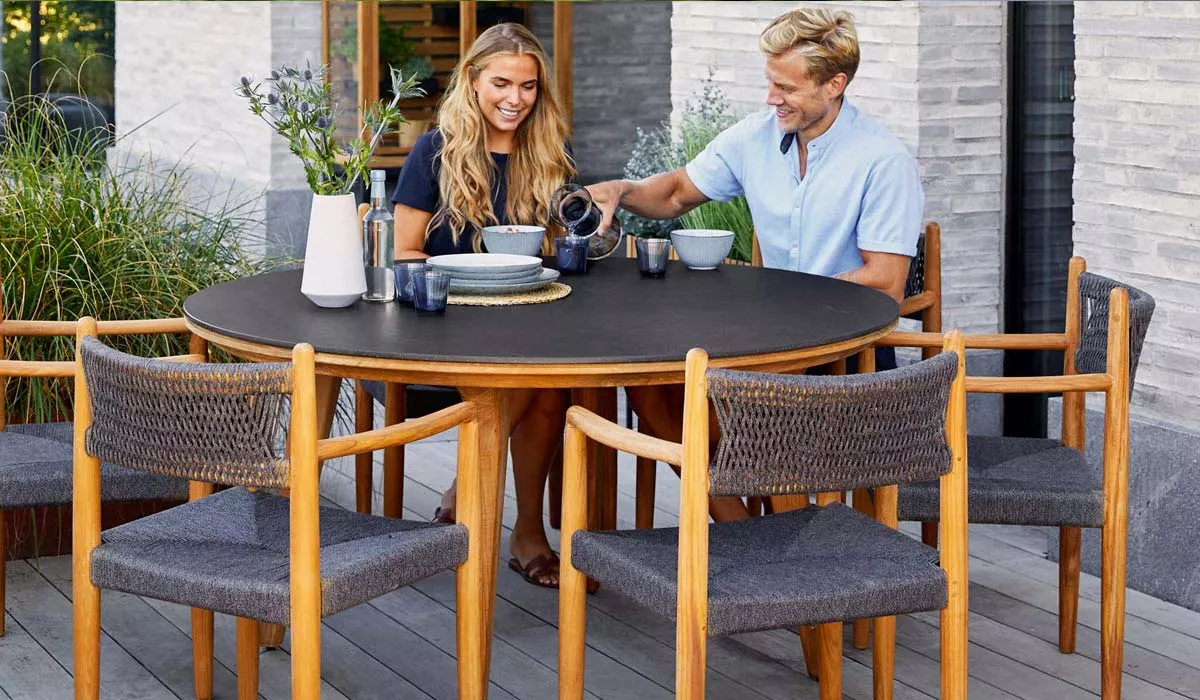 Zwei Personen sitzen an einem runden Cane-line Essgruppen-Tisch aus Holz mit grauen Stühlen auf einer Terrasse vor einer Steinwand.