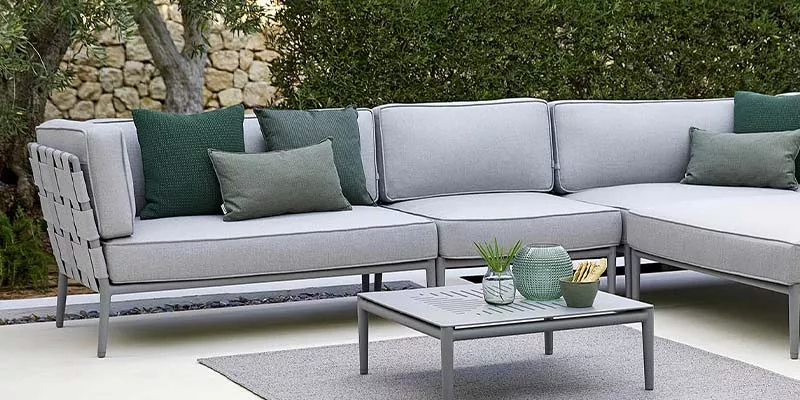 Cane-line Conic Loungegruppe in Grau auf einer Terrasse mit grünen Kissen, umgeben von Hecken, auf einem hellen Boden mit passendem Tisch.