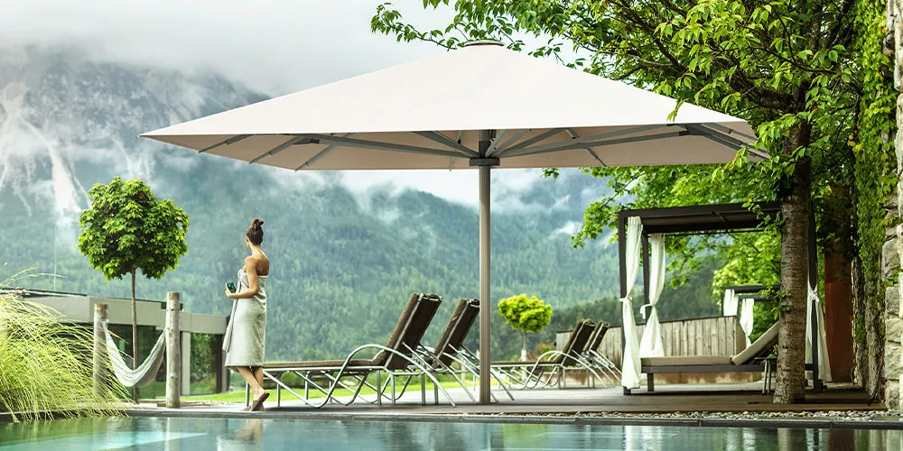 Frau in Garten mit großem, beigem Sonnenschirm von Villa Schmidt, umgeben von Liegestühlen und Bäumen, vor Bergkulisse.
