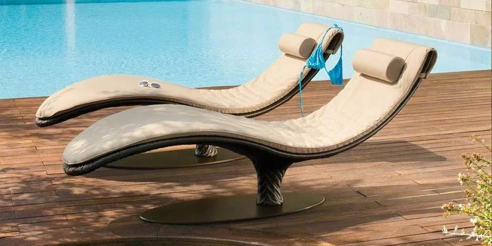 Zwei beige Roberti Caribe Outdoor-Liegen auf Holzterrasse vor einem Pool, mit geschwungenem Design und Kopfstützen.