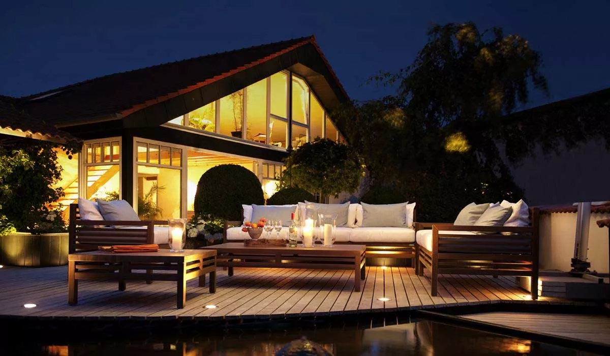 Herrenhaus Cubic Gartenmöbel in dunklem Holz auf beleuchtetem Terrassenboden vor modernem Haus bei Nacht, stimmungsvolle Atmosphäre.