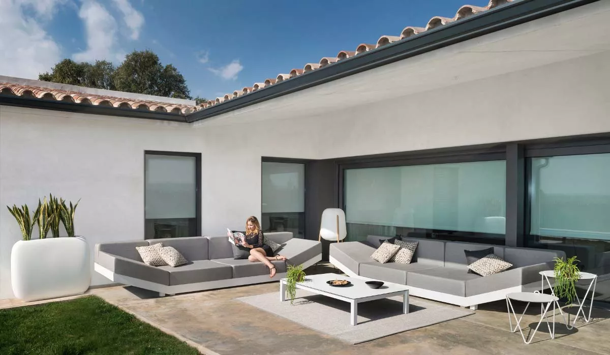 Frau entspannt auf grauem Calma Loungesofa in modernem Garten mit weißem Tisch und Pflanzen, sonniger Tag.