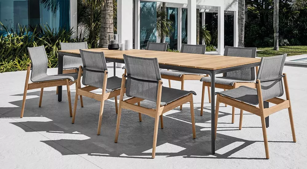 Gloster Sway Gartenmöbel Essgruppe aus Holz und grauem Stoff auf einer Terrasse vor einem modernen Haus mit Palmen im Hintergrund.